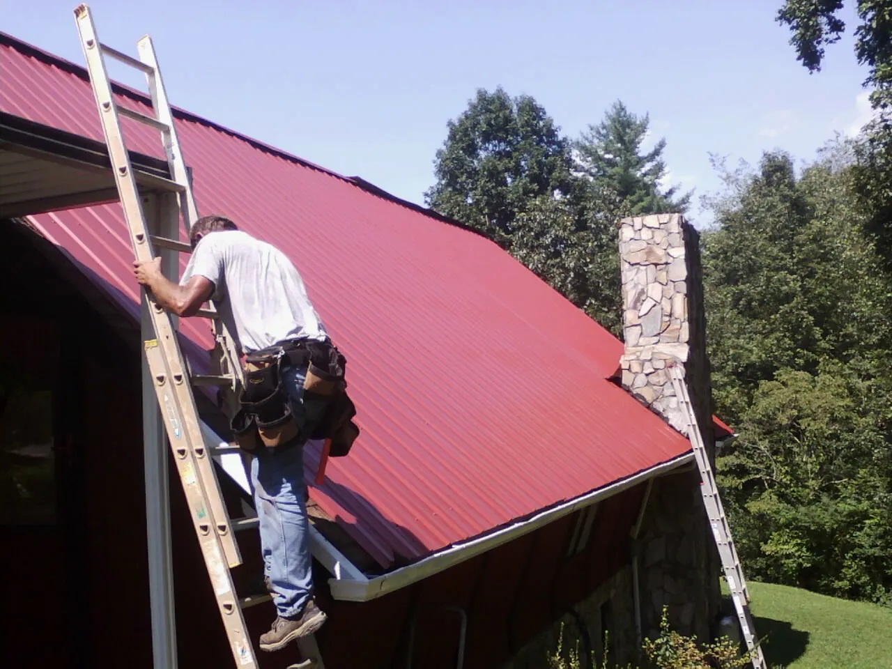 Roofer on a ladder beside a red metal roof and stone chimney — Soffit & Fascia Repair by Roofing Brothers in Suches